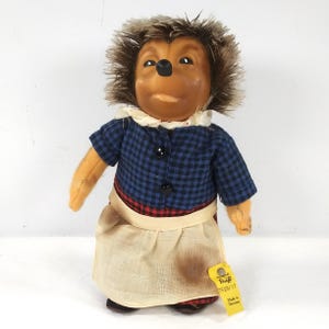 May include: A vintage plush hedgehog doll wearing a blue and black plaid shirt, a red and black plaid apron, and a white skirt. The doll has a wooden head with a painted face and brown hair. The doll has a tag that reads "Original Steiff 1628/17 Made in Germany".