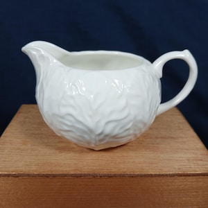 May include: A white ceramic creamer featuring a textured surface that mimics the appearance of overlapping cabbage leaves. The vessel includes a curved pouring spout and a small handle on the side, finished with a smooth, glossy glaze.