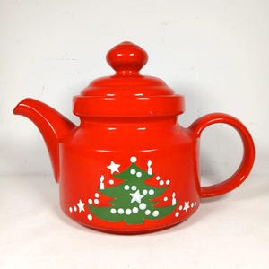 May include: A bright red ceramic teapot with a matching lid, featuring a green Christmas tree graphic decorated with white dots, stars, and candle shapes on the front. The teapot has a curved handle and a pointed spout, with a glossy finish.