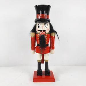 Könnte beinhalten: Ein traditioneller Nussknacker aus Holz mit einer rot-goldenen Uniform, schwarzen Haaren und einem schwarzen Zylinder. Der Nussknacker steht auf einem roten Sockel.