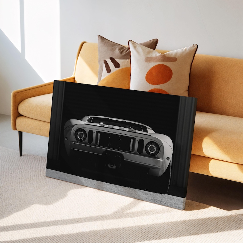 May include: A black and white framed canvas print of a vintage sports car. The car is white with round headlights and a black grille. The print is leaning against a yellow sofa with decorative pillows.