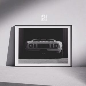 May include: Black and white framed print of a classic sports car, viewed from the rear. The car is white with a central stripe. The print is displayed in a garage setting, with the car's details highlighted by the lighting.
