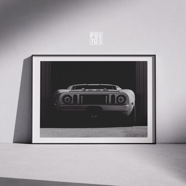 May include: Black and white framed print of a classic sports car, viewed from the rear. The car is white with a central stripe. The print is displayed in a garage setting, with the car's details highlighted by the lighting.