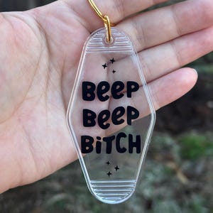 May include: Clear acrylic keychain with a gold-colored keyring. The keychain has the words "BEEP BEEP BITCH" in black lettering, with four small star graphics. The keychain is in a motel key shape.
