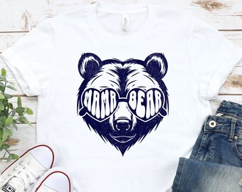 以下が含まれることがあります： 白いTシャツに、ハート型のサングラスをかけて「mama bear」と書かれた青いクマが描かれています。Tシャツは白い木製の表面に置かれ、白いスニーカーと青いジーンズが置かれています。