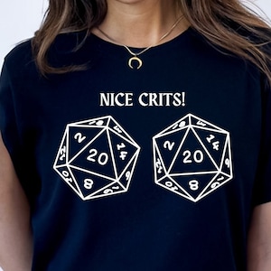 以下が含まれることがあります： 「NICE CRITS!」という白い文字と、20面ダイスの白い画像が2つ入った黒いTシャツ。ダイスには20の数字が表示されています。