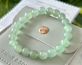 Può includere: Un braccialetto di avventurina verde in una pochette verde chiaro con il testo "Stone & Sonder" sul davanti. La pochette è appoggiata su una superficie bianca.
