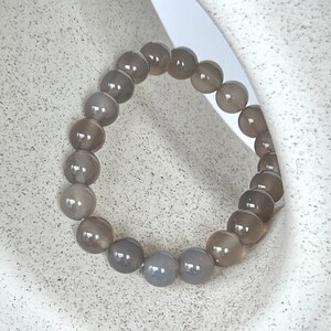 May include: A grey beaded bracelet made of round, smooth stones. The bracelet is lying on a white textured surface.