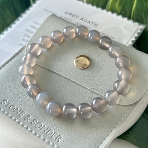 May include: Grey agate bracelet in a white pouch with the text "STONE & SONDER" on it. The pouch is open and the bracelet is visible. The bracelet is made of grey agate beads.