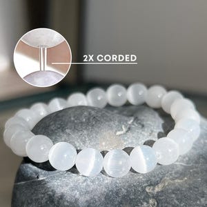 May include: White gemstone bracelet with 2x corded closure. The bracelet is made of round, smooth beads and is shown on a grey stone surface.