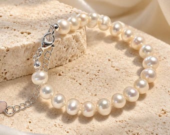 May include: A bracelet composed of round, off-white freshwater pearls strung together. The piece features a silver-toned metal lobster claw clasp, an adjustable extender chain, and a small heart-shaped charm at the end of the chain.