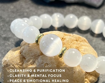 May include: White gemstone bracelet with gold beads. The bracelet is on a light brown rock. The text on the image reads: "Cleansing & Purification, Clarity & Mental Focus, Peace & Emotional Healing."