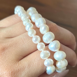 K&ouml;nnte beinhalten: Ein zweireihiges Armband aus wei&szlig;en, unregelm&auml;&szlig;ig geformten S&uuml;&szlig;wasserperlen. Die Perlen variieren in ihrer Gr&ouml;&szlig;e und weisen eine gl&auml;nzende, schimmernde Oberfl&auml;che auf, die zu einem flexiblen, mehrlagigen Schmuckst&uuml;ck aneinandergereiht sind.