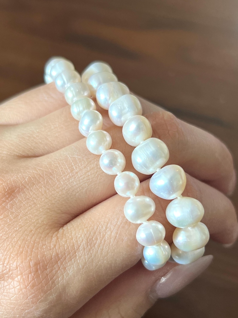 K&ouml;nnte beinhalten: Ein zweireihiges Armband aus wei&szlig;en, unregelm&auml;&szlig;ig geformten S&uuml;&szlig;wasserperlen. Die Perlen variieren in ihrer Gr&ouml;&szlig;e und weisen eine gl&auml;nzende, schimmernde Oberfl&auml;che auf, die zu einem flexiblen, mehrlagigen Schmuckst&uuml;ck aneinandergereiht sind.