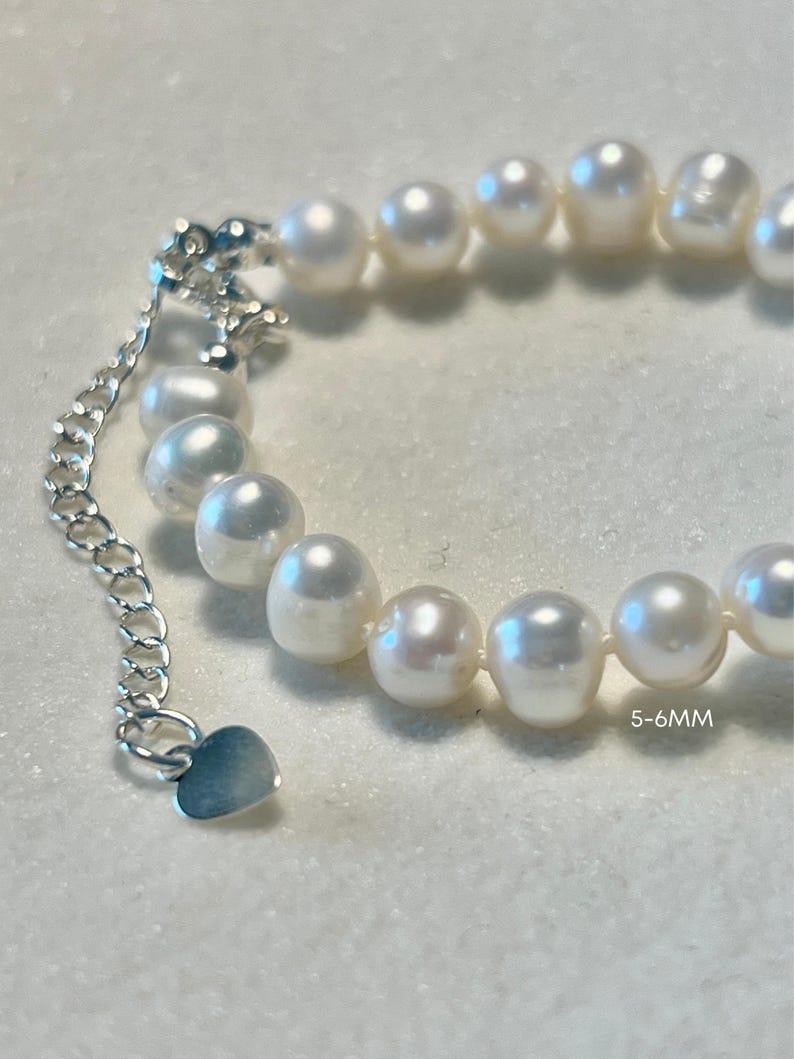 K&ouml;nnte beinhalten: Ein Armband aus wei&szlig;en, gl&auml;nzenden, fast runden Perlen mit einem Durchmesser von 5-6mm. Das Schmuckst&uuml;ck verf&uuml;gt &uuml;ber eine silberfarbene Metall-Verl&auml;ngerungskette, die mit einem kleinen herzf&ouml;rmigen Anh&auml;nger am Ende versehen ist.