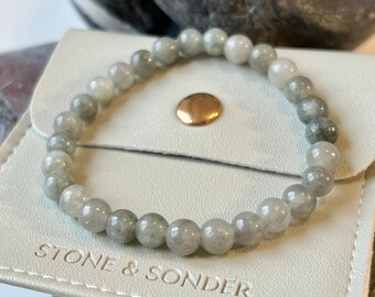 May include: A gray gemstone bracelet with a light gray and white color gradient. The bracelet is on a white leather pouch with the text "Stone & Sonder" embossed on it.