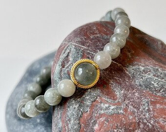 May include: A gray gemstone bracelet with a gold-toned accent bead. The bracelet is resting on a gray and red rock.