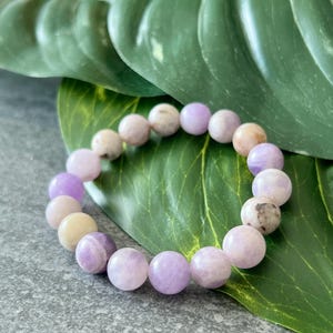 Puede incluir: Un brazalete de piedras preciosas de color p&uacute;rpura y blanco con cuentas redondas. El brazalete est&aacute; sobre un fondo de hoja verde.
