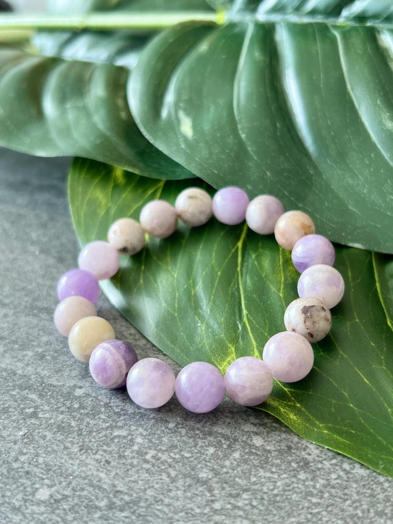 Puede incluir: Un brazalete de piedras preciosas de color p&uacute;rpura y blanco con cuentas redondas. El brazalete est&aacute; sobre un fondo de hoja verde.