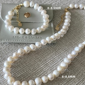 K&ouml;nnte beinhalten: Ein Set aus wei&szlig;em Perlenarmband und Perlenkette mit goldfarbenen Metallverschl&uuml;ssen und herzf&ouml;rmigen Anh&auml;ngern. Die Perlen haben einen Durchmesser von 8-8,5 mm. Das Set liegt auf einem grauen Beutel mit dem Aufdruck STONE & SONDER.
