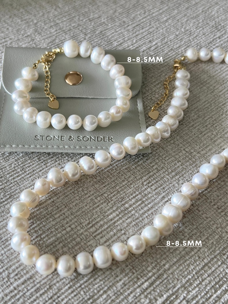 K&ouml;nnte beinhalten: Ein Set aus wei&szlig;em Perlenarmband und Perlenkette mit goldfarbenen Metallverschl&uuml;ssen und herzf&ouml;rmigen Anh&auml;ngern. Die Perlen haben einen Durchmesser von 8-8,5 mm. Das Set liegt auf einem grauen Beutel mit dem Aufdruck STONE & SONDER.