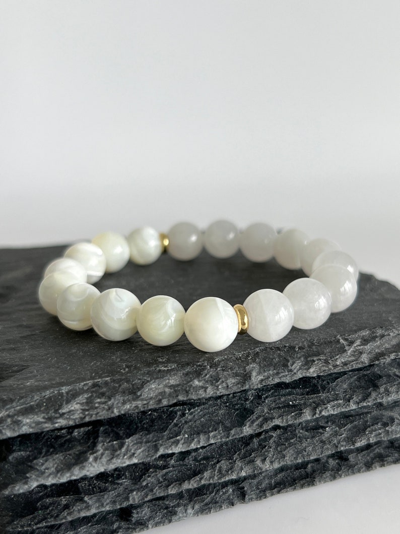 May include: White gemstone bracelet with gold accents. The bracelet is made of round beads and is strung on an elastic cord.