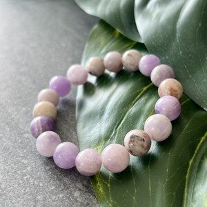 Puede incluir: Un brazalete de piedras preciosas de color p&uacute;rpura y blanco con cuentas redondas. El brazalete est&aacute; sobre una hoja verde.