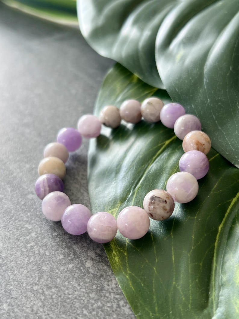 Puede incluir: Un brazalete de piedras preciosas de color p&uacute;rpura y blanco con cuentas redondas. El brazalete est&aacute; sobre una hoja verde.