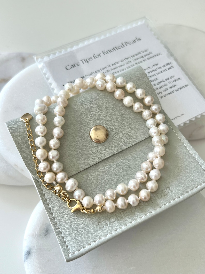K&ouml;nnte beinhalten: Eine wei&szlig;e Perlenkette mit goldfarbener Metallverl&auml;ngerungskette und Verschluss, liegend auf einem hellgrauen quadratischen Beutel mit goldenem Druckknopf und dem gepr&auml;gten Text STONE & SENDER, begleitet von einer Karte Care Tips for Knotted Pearls.