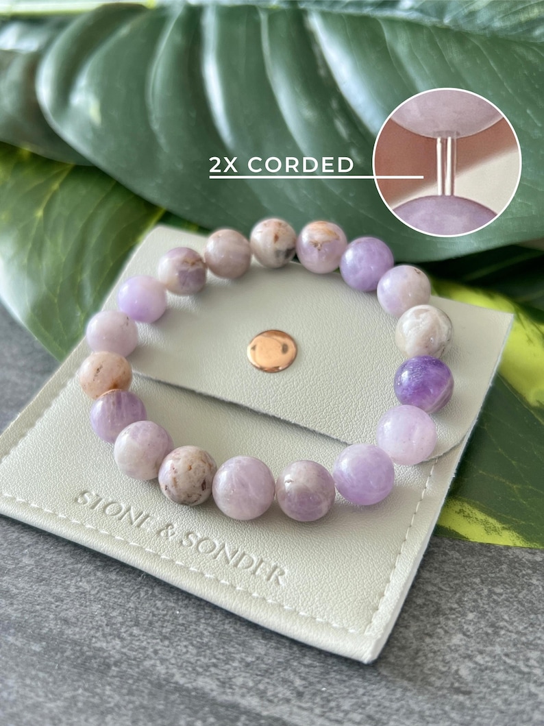 Puede incluir: Un brazalete de piedras preciosas de amatista p&uacute;rpura con cierre de doble cord&oacute;n. El brazalete se muestra en una bolsa de cuero blanco con el texto "Stone & Sonder" impreso en ella. La imagen tambi&eacute;n incluye el texto "2X Corded".