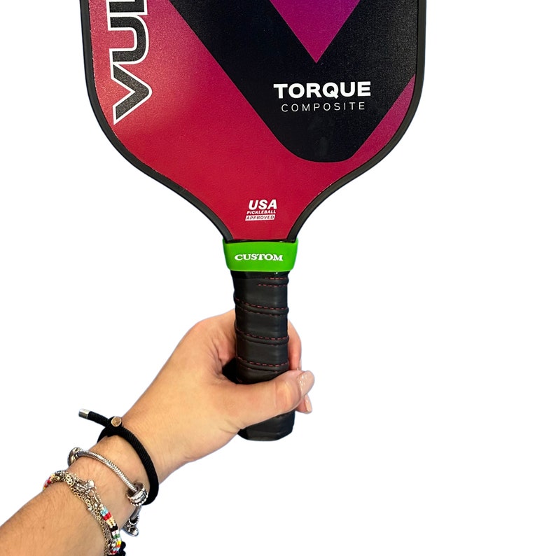 May include: A Vulcan Torque Composite pickleball paddle with a black, red, and purple gradient face. The paddle features the text VULCAN, TORQUE COMPOSITE, and USA PICKLEBALL APPROVED. It has a black handle with red stitching and a bright green band labeled CUSTOM.