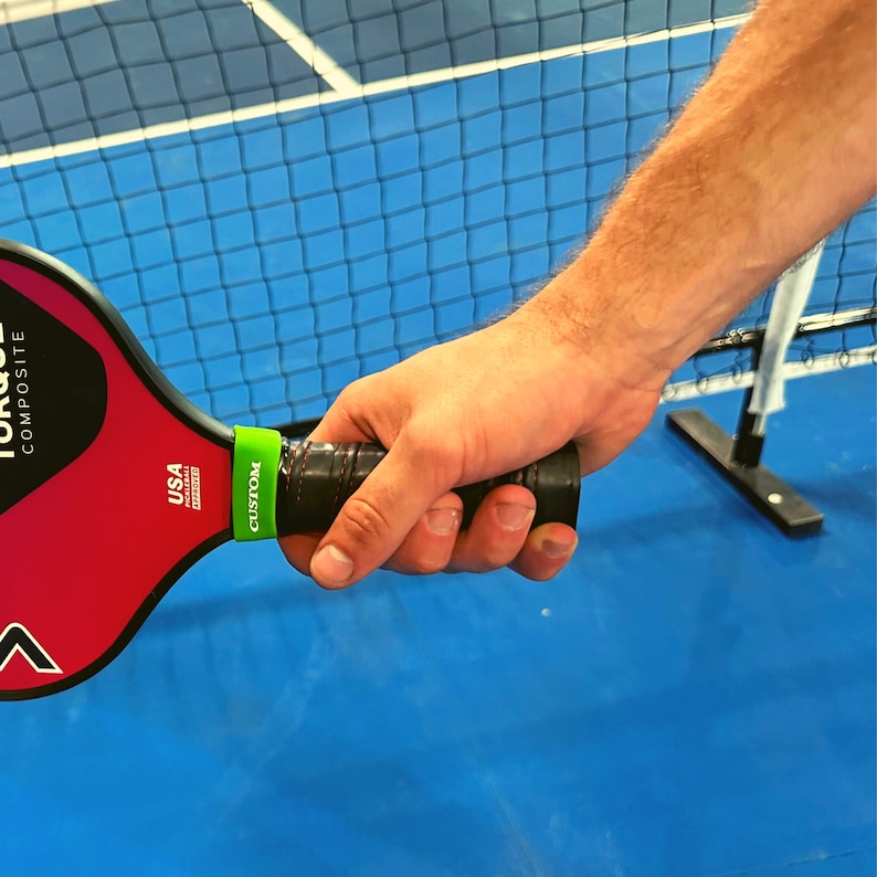 May include: A red and black composite pickleball paddle featuring the text TORQUE COMPOSITE and USA PICKLEBALL APPROVED. The handle is wrapped in black tape with red stitching and includes a bright green band with the word CUSTOM printed on it.