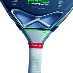 May include: A black and silver padel racket featuring the text AT10 GENIUS, AGUSTIN TAPIA, LUXURY SERIES, HR3 CORE, and CARBON 12K. The handle is wrapped in white grip tape with a red band featuring the white text CARLOS near the throat.