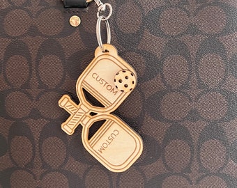 May include: A wooden pickleball paddle keychain with a small ball charm. The paddle is engraved with the word "CUSTOM" on both sides.