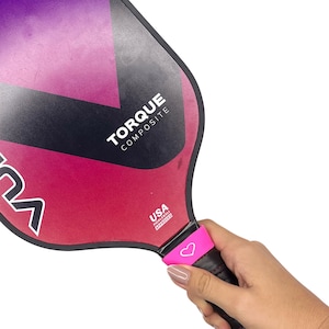 May include: A pickleball paddle with a black, purple, and pink gradient surface. The paddle features the text TORQUE COMPOSITE and a USA PICKLEBALL APPROVED logo. A bright pink rubber band with a white heart icon is wrapped around the black handle.
