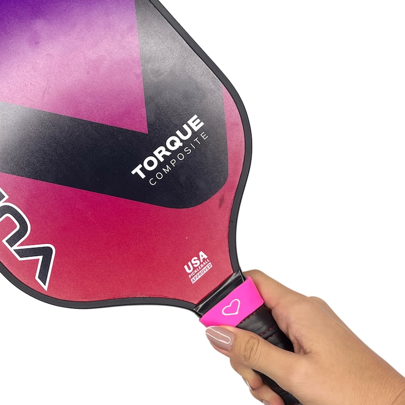 May include: A pickleball paddle with a black, purple, and pink gradient surface. The paddle features the text TORQUE COMPOSITE and a USA PICKLEBALL APPROVED logo. A bright pink rubber band with a white heart icon is wrapped around the black handle.