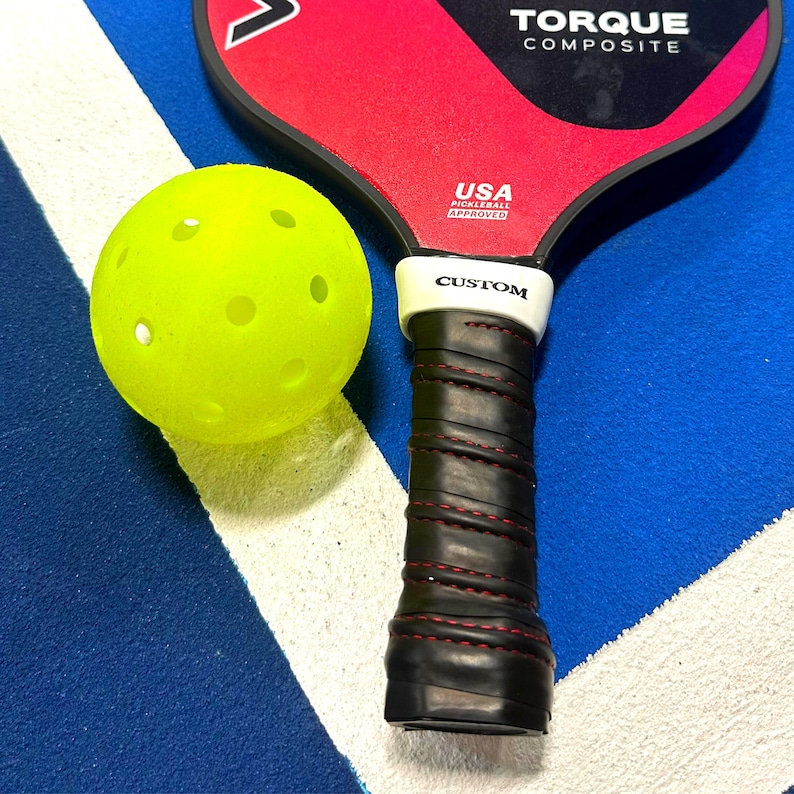 May include: A neon yellow pickleball with circular perforations positioned next to a black and red pickleball paddle. The paddle features the text TORQUE COMPOSITE, USA PICKLEBALL APPROVED, and CUSTOM on the white grip collar with black tape.