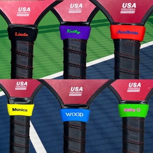 May include: Six pickleball paddle handles featuring black grips with red stitching and personalized silicone bands in black, purple, red, yellow, blue, and green. Each band displays a name and the red paddle face shows USA Pickleball Approved text.