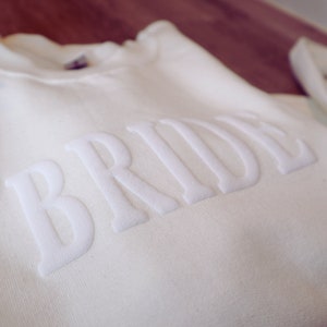 May include: White sweatshirt with the word "BRIDE" in raised white lettering.