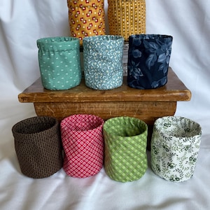 May include: A collection of eight fabric containers in various patterns and colors. The containers are cylindrical with a gathered top. Patterns include floral, geometric, and leaf designs in shades of yellow, blue, green, red, and brown.
