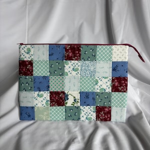 May include: A patchwork zippered pouch featuring a variety of floral and geometric patterns in shades of blue, green, and burgundy. The pouch has a burgundy zipper and pull, and is likely used for carrying small items.