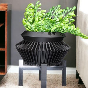 Puede incluir: Una maceta negra geométrica con un diseño decorativo en forma de abanico, que contiene un follaje verde vibrante. La maceta se asienta sobre un soporte negro a juego, creando una estética moderna. La maceta tiene aproximadamente 30 cm de diámetro.