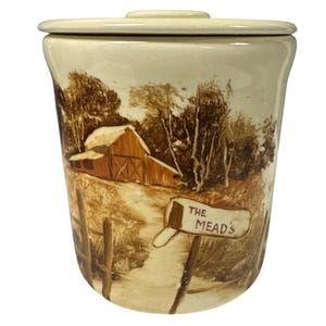 May include: A cream-colored ceramic canister with a lid, featuring a rural landscape scene. The artwork depicts a barn, trees, and a mailbox with "The Mead's" written on it. The canister has a slightly flared shape.