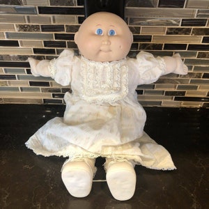 May include: A Cabbage Patch Kid doll wearing a white dress with lace trim and white shoes. The doll has a bald head, blue eyes, and a rosy complexion.