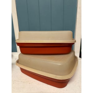 May include: Two vintage food storage containers. The containers are rectangular with a red base and a beige lid. The lids have a slightly textured surface. The containers are stacked on top of each other.