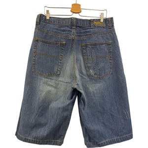 May include: Blue denim jean shorts with a hand design on the back pocket. The shorts have a distressed look with fading and a small logo on the back. The shorts are hanging on a wooden hanger.