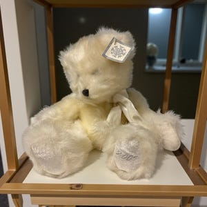 Op de afbeelding: Een crèmekleurige teddybeer met een zwarte neus en een wit lint om zijn nek. De beer heeft een label met een sneeuwvlokontwerp en het nummer "2000" geborduurd op zijn voet. De beer zit op een witte plank.