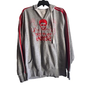 May include: A gray hooded sweatshirt with red accents. The front features a red graphic of a person with sunglasses and the text "NOT IN MY HOUSE." Red stripes run down the sleeves.