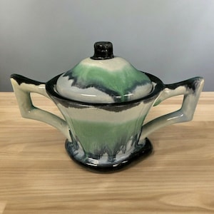 May include: A green, white, and black ceramic sugar bowl with a lid. The bowl has a unique, abstract design and two handles.