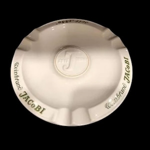May include: White ceramic ashtray with a gold design. The ashtray has the words "Weinbrand JACOBI" written around the edge and a gold "J" logo in the centre.